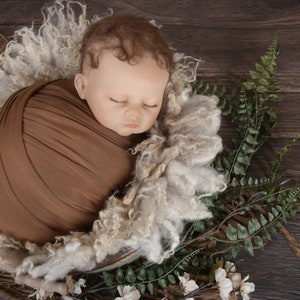 May include: A newborn baby is swaddled in a brown fabric wrap and placed in a white fluffy nest. The baby is sleeping peacefully with its eyes closed. The nest is surrounded by green ferns and white flowers.