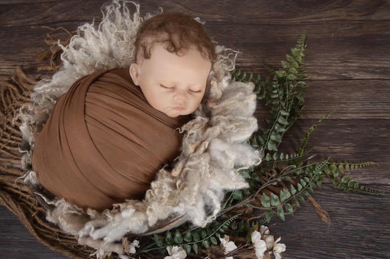 May include: A newborn baby is swaddled in a brown fabric wrap and placed in a white fluffy nest. The baby is sleeping peacefully with its eyes closed. The nest is surrounded by green ferns and white flowers.