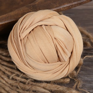 May include: A beige stretchy fabric swaddle for newborn photography. The fabric is tightly wound into a ball shape.
