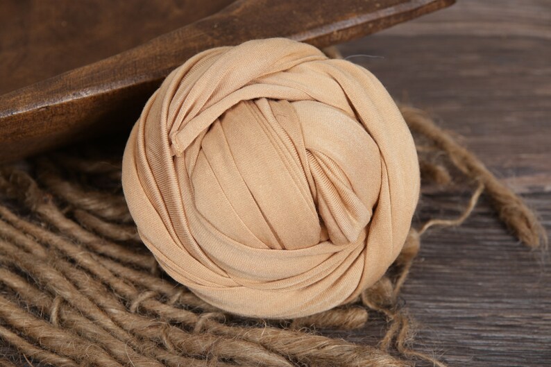 May include: A beige stretchy fabric swaddle for newborn photography. The fabric is tightly wound into a ball shape.