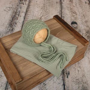 May include: A sage green knitted baby bonnet with a diamond pattern and matching ribbed fabric. The bonnet has long ties and is placed on a wooden prop box. The background is a rustic, light-colored wood.