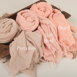 May include: Three different shades of pink fabric swaddles, labeled "Porcelain", "Soft Shell", and "Peach". The fabric is soft and textured, with a slightly crinkled appearance.