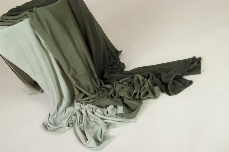 May include: A pile of fabric in shades of green and white. The fabric is draped over a wooden box and flows down onto a white surface.