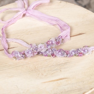May include: A light purple floral headband and matching tieback, featuring delicate fabric flowers and pearl accents. The set is made of soft, flowing fabric and is designed for newborns or infants. The items are displayed on a light wooden surface.