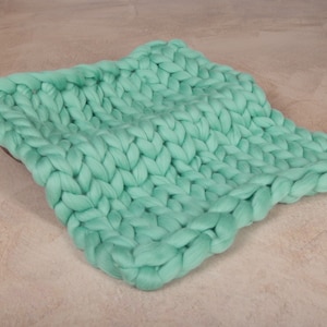 May include: A large, chunky knit blanket in a light green color. The blanket is made of thick yarn and has a soft, fluffy texture.