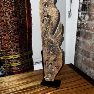 May include: A tall, vertical wooden carving featuring an intricate, textured relief of a mythical serpentine creature with scales and wings. The sculpture is finished in a weathered brown tone and is mounted on a simple, rectangular black metal stand.