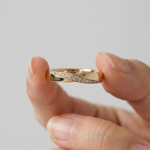 Könnte beinhalten: Ein goldener Ring mit einer diagonalen Fassung, die mit kleinen klaren Steinen besetzt ist. Das Metall hat eine polierte Oberfläche und das Design enthält ein dezentes Dreh- oder Kreuzungselement im vorderen Bereich des Rings für eine elegante Optik.
