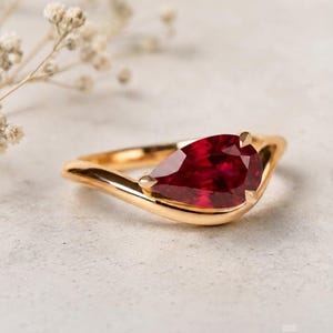 Puede incluir: Un anillo de oro con una piedra preciosa de color rojo intenso en forma de pera, montada en una banda lisa y pulida. El engaste sujeta la piedra de forma segura con pequeñas garras, creando un aspecto elegante y continuo en la banda de metal.