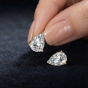 Puede incluir: Un par de pendientes en forma de pera con piedras transparentes facetadas engastadas en garras de metal dorado. Un pendiente se sostiene entre los dedos, mientras que el otro descansa cerca, mostrando el corte brillante de las piedras.