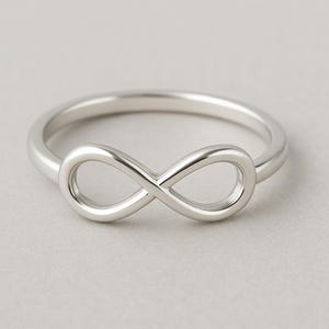 Può includere: Un anello color argento lucido caratterizzato da un design centrale a forma di simbolo dell'infinito. La fascia è liscia e arrotondata e si collega alla forma a ciclo continuo del motivo dell'infinito, integrato nella parte anteriore dell'anello.