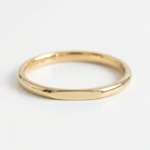 Può includere: Un anello in oro giallo lucido caratterizzato da una fascia liscia e arrotondata con una sezione piatta rettangolare sulla parte anteriore. Il metallo ha una finitura riflettente ad alta lucentezza che cattura la luce, creando una forma.