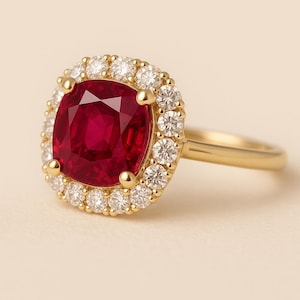 May include: A gold ring featuring a large, cushion-cut, deep red ruby center stone surrounded by a halo of small, round, brilliant-cut white diamonds. The setting is crafted from polished yellow gold, showcasing a classic, elegant design.