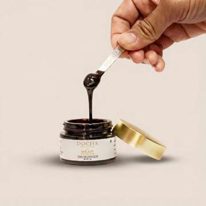 Op de afbeelding: Een klein, helder glazen potje met een gouden deksel en een donkerbruine substantie erin. Een hand houdt een kleine zilveren lepel vast, waarmee de substantie in het potje wordt gedruppeld. Het potje heeft een wit etiket met het woord "DOCHA".
