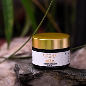 Op de afbeelding: Een pot DOCHA Ayurveda Shilajit, een resin-royal blend, staat op een stenen oppervlak. De pot heeft een goudkleurige deksel en een wit etiket met zwarte tekst. De achtergrond toont groen gebladerte.