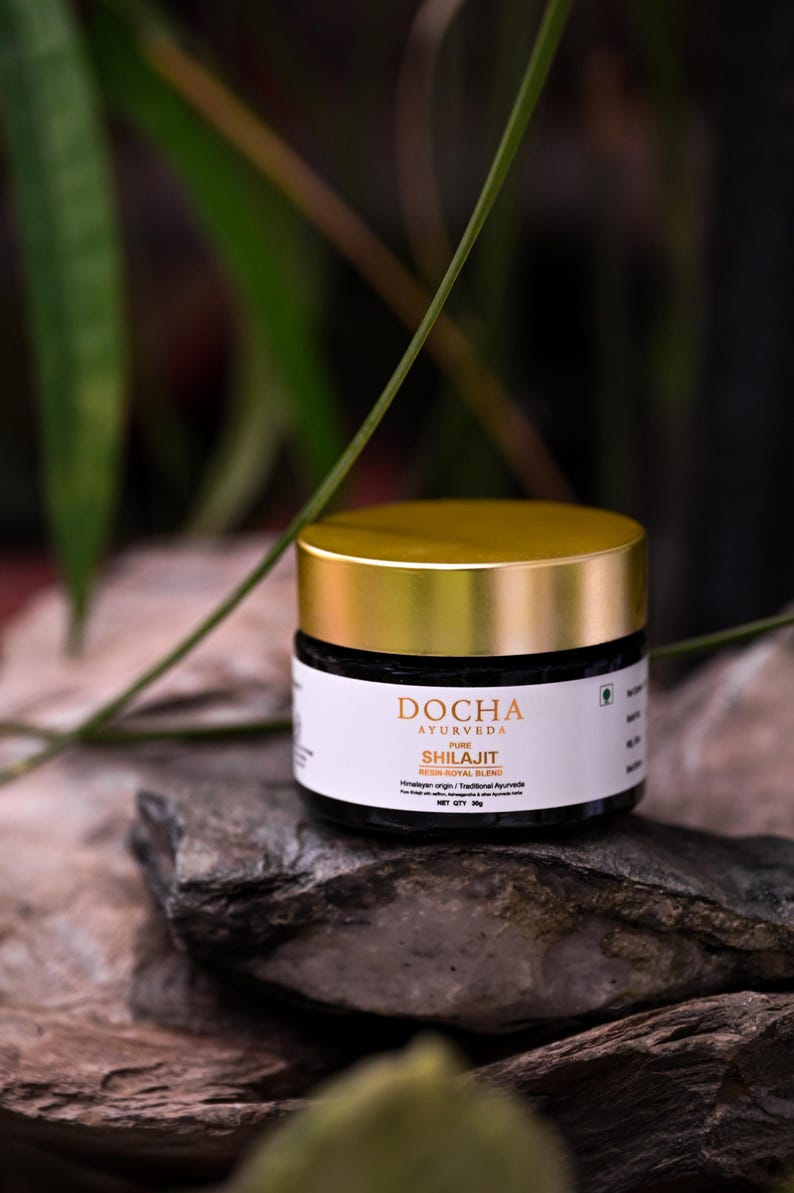 Op de afbeelding: Een pot DOCHA Ayurveda Shilajit, een resin-royal blend, staat op een stenen oppervlak. De pot heeft een goudkleurige deksel en een wit etiket met zwarte tekst. De achtergrond toont groen gebladerte.