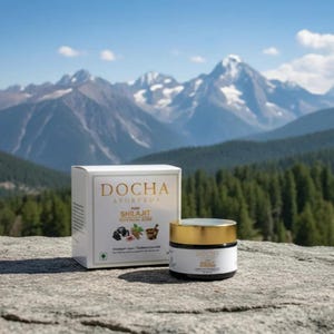 Op de afbeelding: Een witte doos en een klein potje huidverzorgingsproducten worden getoond op een rotsachtig oppervlak, met een schilderachtige bergachtergrond. De doos heeft gouden letters met de tekst "DOCHA AYURVEDA". De pot heeft een gouden deksel en een wit etiket.
