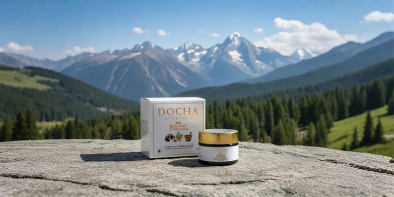 Op de afbeelding: Een witte doos en een klein potje huidverzorgingsproducten worden getoond op een rotsachtig oppervlak, met een schilderachtige bergachtergrond. De doos heeft gouden letters met de tekst "DOCHA AYURVEDA". De pot heeft een gouden deksel en een wit etiket.