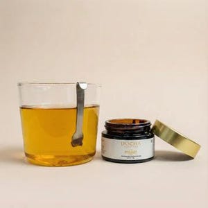 Op de afbeelding: Een glas amberkleurige thee met een roestvrijstalen thee-ei. Naast het glas staat een klein potje met een gouden deksel en een etiket met "DOCHA" en "SHIKAI".