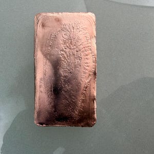 Könnte beinhalten: Ein rechteckiger Kupferbarren mit metallisch rotbrauner Oberfläche. Die Mitte weist ein ausgeprägtes, texturiertes, farnartiges Kristallmuster auf, während die Kanten glatt und leicht abgerundet mit einem polierten Glanz erscheinen.