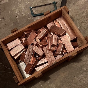 Könnte beinhalten: Eine rechteckige Holzkiste gefüllt mit zahlreichen metallischen, kupferfarbenen Barren unterschiedlicher Größe und Textur. Eine kleine Metallsäge mit blauem Griff liegt auf dem Rand der Kiste, nahe der oberen rechten Ecke des Behälters.