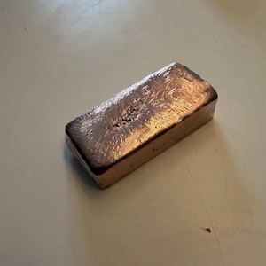 Könnte beinhalten: Ein rechteckiger, kupferfarbener Metallbarren mit einer texturierten, unebenen Oberfläche. Das Objekt hat abgerundete Kanten und einen metallischen Glanz, der sichtbare Anzeichen von Guss oder Formgebung mit einer rauen Oberflächenstruktur aufweist.