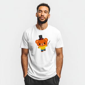 May include: White t-shirt featuring a cartoon bear wearing a top hat and holding a wand. The bear is orange with a yellow shirt, blue pants, and a black top hat. The design is centered on the shirt.