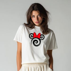 May include: A white short-sleeved t-shirt featuring a graphic print on the chest. The design consists of a black spiral-shaped silhouette resembling a mouse head, accented with a bright red bow positioned between the two circular ears.