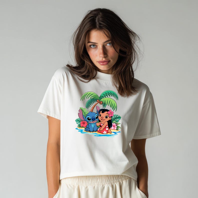 May include: Off-white t-shirt featuring a colorful graphic of Lilo and Stitch on a tropical beach scene. The design includes a blue alien, a girl in a red dress, palm trees, and flowers. The shirt has a crew neck and short sleeves.
