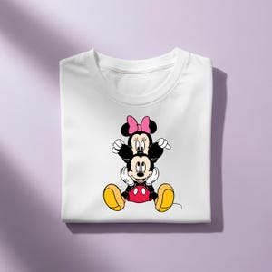 May include: A white t-shirt featuring a graphic print of Minnie Mouse sitting on top of Mickey Mouse. Minnie wears a pink bow, while Mickey is shown with his signature red shorts and yellow shoes. The characters are stacked vertically in the center.