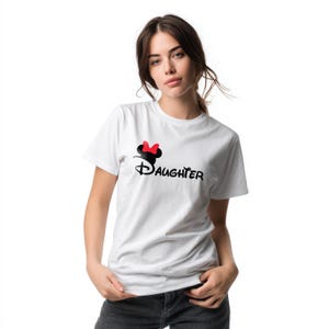 May include: A white short-sleeved t-shirt featuring a black graphic of a mouse ear silhouette with a red bow above the word Daughter written in a stylized black font across the chest area of the garment.