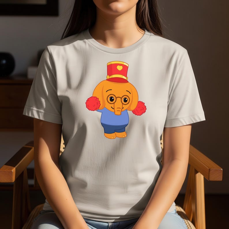 May include: A light gray t-shirt featuring a cartoon elephant character. The elephant has orange skin, round glasses, a red and gold hat, and red pom-poms. The elephant is wearing a blue shirt and shorts.