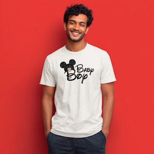 May include: A light gray short-sleeved t-shirt featuring a black graphic of a mouse silhouette with a small colorful pacifier in the center, accompanied by the black text Baby Boy written in a stylized script font across the chest area.