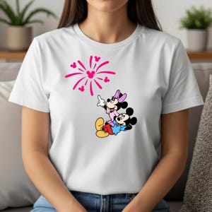 May include: A white short-sleeved t-shirt featuring a graphic print of Mickey and Minnie Mouse looking up at a pink firework display. The firework design incorporates small Mickey Mouse head silhouettes within the radiating lines of the pattern.