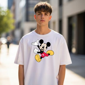 May include: A white short-sleeved t-shirt featuring a graphic print of Mickey Mouse in a dynamic pose. The character is depicted with his signature black ears, red shorts, yellow shoes, and white gloves, centered on the front of the garment.