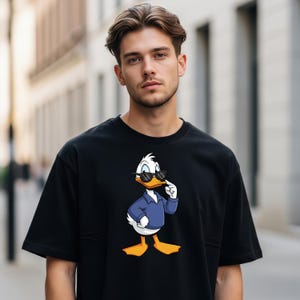 May include: A black short-sleeved t-shirt featuring a graphic print of a white duck wearing a blue collared shirt and black sunglasses. The duck is standing with one wing raised toward its face in a pose that mimics a thoughtful expression.