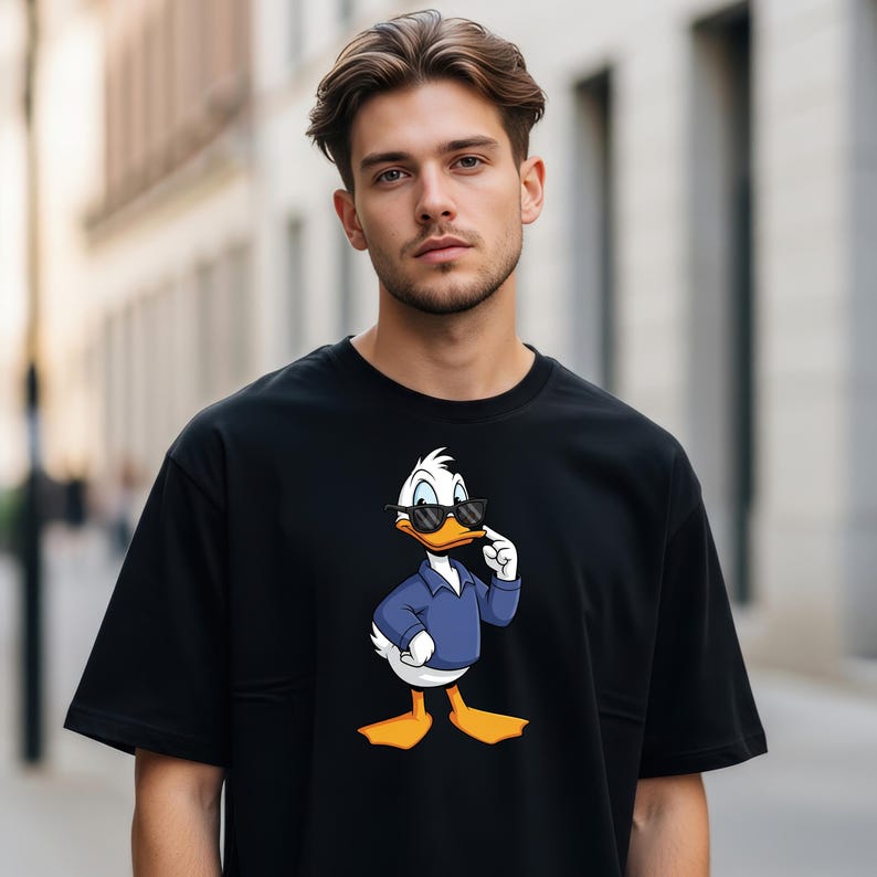 May include: A black short-sleeved t-shirt featuring a graphic print of a white duck wearing a blue collared shirt and black sunglasses. The duck is standing with one wing raised toward its face in a pose that mimics a thoughtful expression.