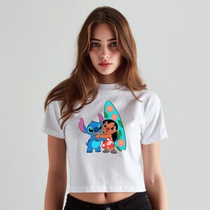 May include: White crop top featuring a colorful graphic of Stitch and Lilo hugging, with a surfboard. The surfboard is teal with orange flower accents. The characters are in a cartoon style.