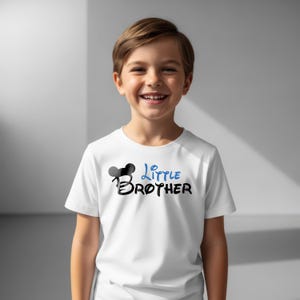 May include: A white short-sleeved t-shirt featuring a graphic design on the chest. The design includes a black silhouette of mouse ears above the word BROTHER in black, with the word Little written in blue script lettering above the word BROTHER.