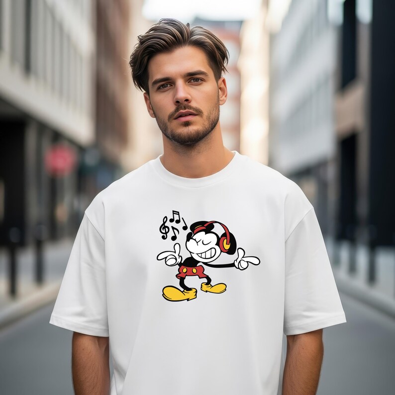 May include: A white short-sleeved t-shirt featuring a graphic print of a cartoon mouse wearing red headphones, red shorts, and yellow shoes, with musical notes floating above the character's head, set against a blurred urban street background.