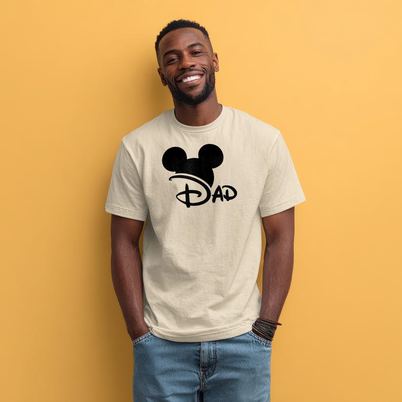 May include: A beige short-sleeved t-shirt featuring a black graphic on the chest. The graphic consists of a stylized mouse ear silhouette positioned above the word DAD, written in a cursive, looping font that mimics a classic script style.