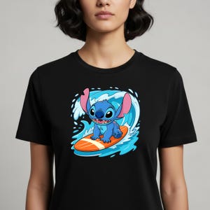 May include: Black t-shirt featuring a cartoon illustration of a blue alien character surfing on an orange surfboard in front of a blue and white wave. The character has large ears and a friendly expression. The shirt is made of a soft material.