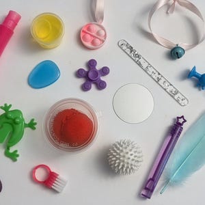 May include: A collection of colorful toys and sensory items, including a pink expandable tube, a green frog, a purple fidget spinner, a blue feather, and a small bell on a pink ribbon. Other items include a small mirror and a container of red playdough.