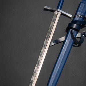 May include: A double-edged sword with a patterned Damascus steel blade, a straight silver crossguard, and a blue leather-wrapped grip. The pommel is circular with a silver inlay. A matching blue leather scabbard with a silver tip is positioned beside it.