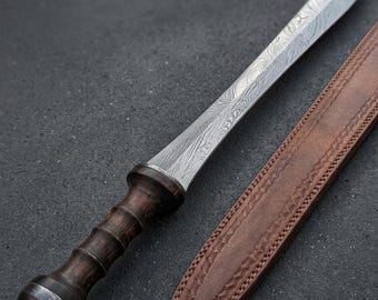 May include: A detailed image of a gladius sword with a patterned steel blade and a dark wooden handle. The sword rests beside a brown leather sheath with decorative stitching. A small, round seal with text is attached.