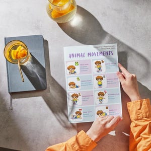 May include: A person holding a printed sheet titled "ANIMAL MOVEMENTS" with illustrations of children performing various exercises. The sheet is on a gray surface next to a blue notebook and a glass of orange-infused water.