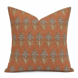 May include: A square throw cushion in a burnt orange hue featuring a repeating block-print style floral pattern. The design consists of symmetrical botanical motifs in muted sage green and mustard yellow, arranged in vertical rows across the fabric.
