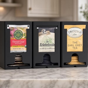 Può includere: Un organizer per bustine di tè nero con tre scomparti verticali. Ogni sezione presenta un'apertura rettangolare che mostra una bustina di tè: Traditional Medicinals Throat Coat, Bio Kräuterfreude e Twinings Earl Grey Tea. L'unità è nero opaco.