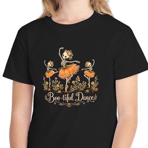 以下が含まれることがあります： 黒の半袖Tシャツ。オレンジ色のカボチャを模したチュチュと花冠を身につけ、秋の花々の間を踊る3体の骸骨のグラフィックが描かれています。骸骨の下には「Boo-tiful Dance」という文字が装飾的なフォントでプリントされています。