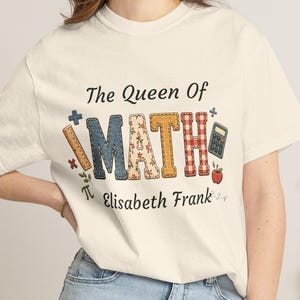 Może przedstawiać: Kremowy t-shirt z krótkim rękawem z napisem The Queen Of MATH wykonanym czcionką w różne wzory. Projekt zawiera linijkę, kalkulator, symbol pi oraz jabłko. Pod głównym tekstem nadrukowane jest imię i nazwisko Elisabeth Frank.