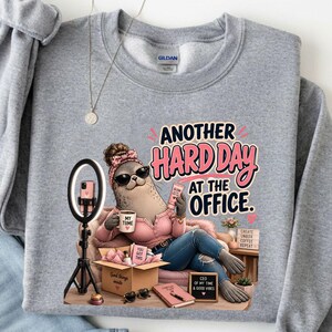 May include: A light gray crewneck sweatshirt featuring a graphic of a seal wearing sunglasses and a pink headband, sitting in an office chair. The text reads Another Hard Day At The Office, with a ring light, a coffee mug, and a cardboard box.